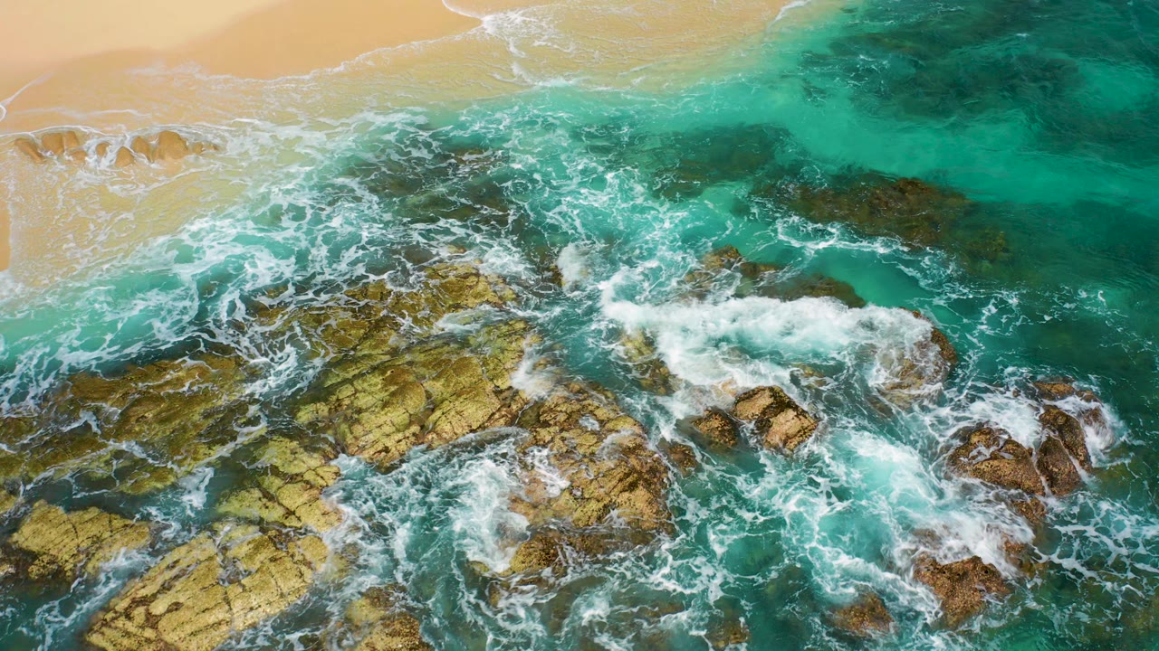 Overhead view in Desert Park Natural Reserve, Baja California Sur, with turquoise waves foaming around brown and yellow rocks below a strip of golden sand, no visible people.