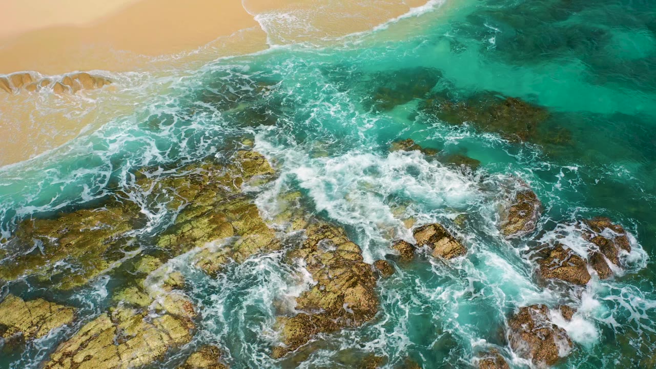 Overhead Desert Park Natural Reserve coastline with a larger white wave wash curling over clustered brown rocks and turquoise water, with sand visible along the top edge and no visible people.