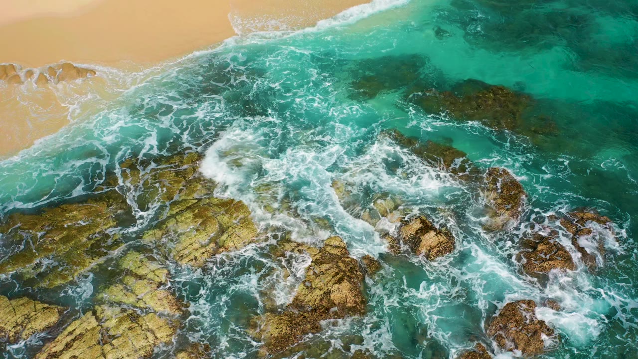 Aerial view over Baja California Sur surf as white foam covers a broad yellow-brown rock ledge in turquoise water, with a small sandy beach edge at the top and no visible people.