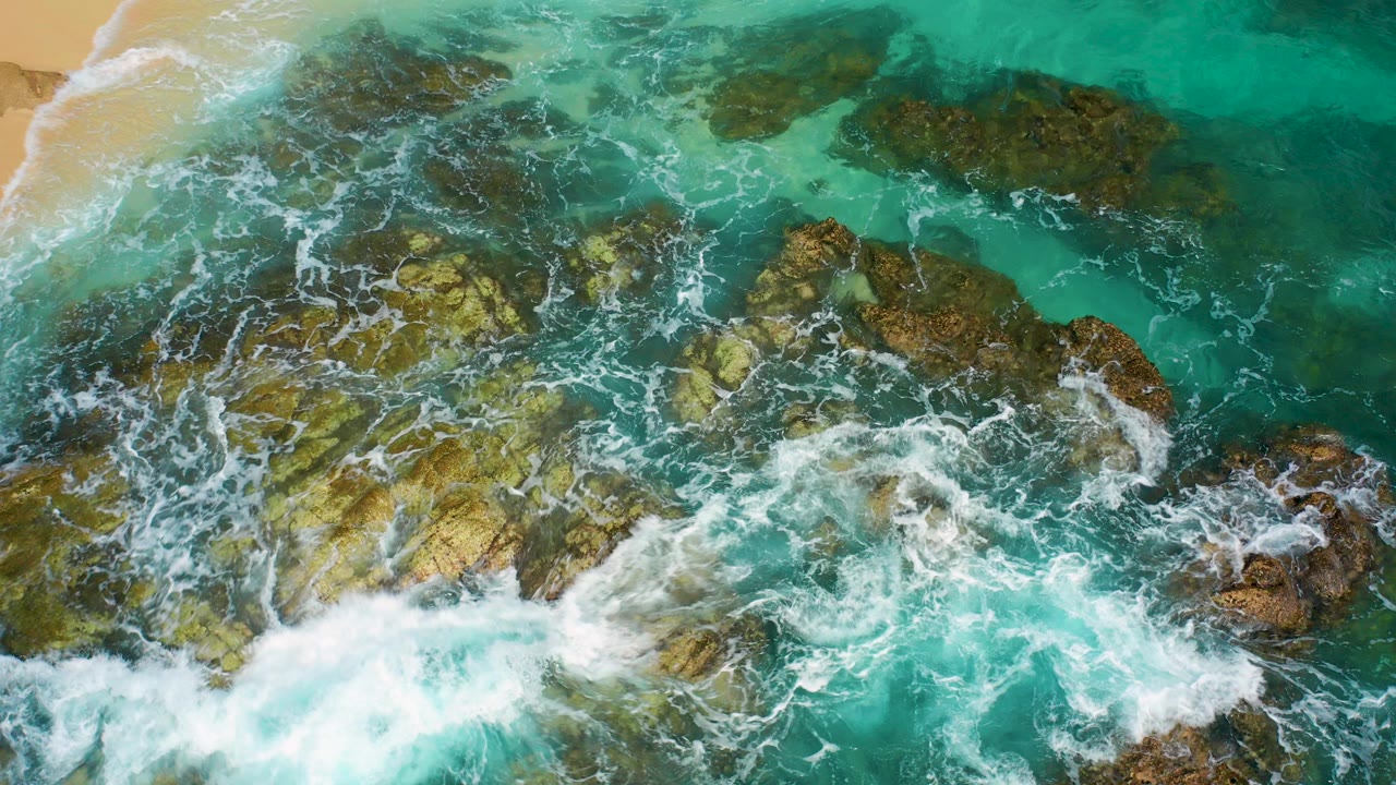 Overhead view of turquoise water and white foam moving across green-brown rocks in Baja California Sur, Mexico, with a tan sand edge at upper left and no visible people.