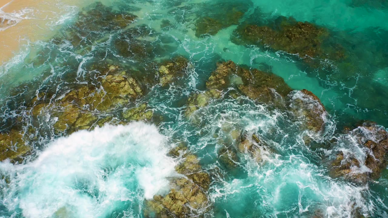 A larger white breaker fills the lower center as turquoise water swirls around exposed brown rocks in Baja California Sur, Mexico, with no visible people.