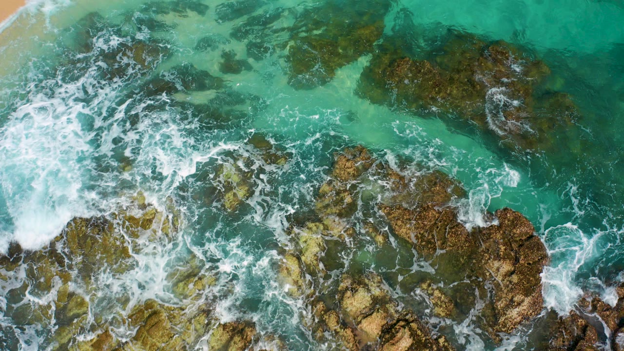 Top-down turquoise surf breaking over brown coastal rocks.
