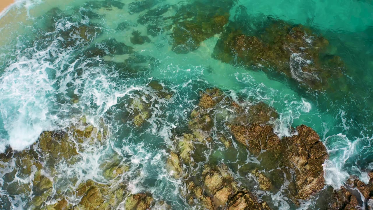 Aerial frame from Desert Park Natural Reserve, Baja California Sur, showing foamy waves spreading over algae-toned rocks in shallow turquoise water with no visible people.
