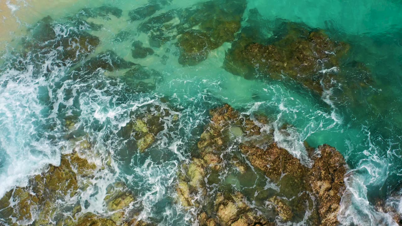 Overhead view in Desert Park Natural Reserve, Baja California Sur, with white surf crossing a dense cluster of brown rocks beneath transparent turquoise water and no visible people.
