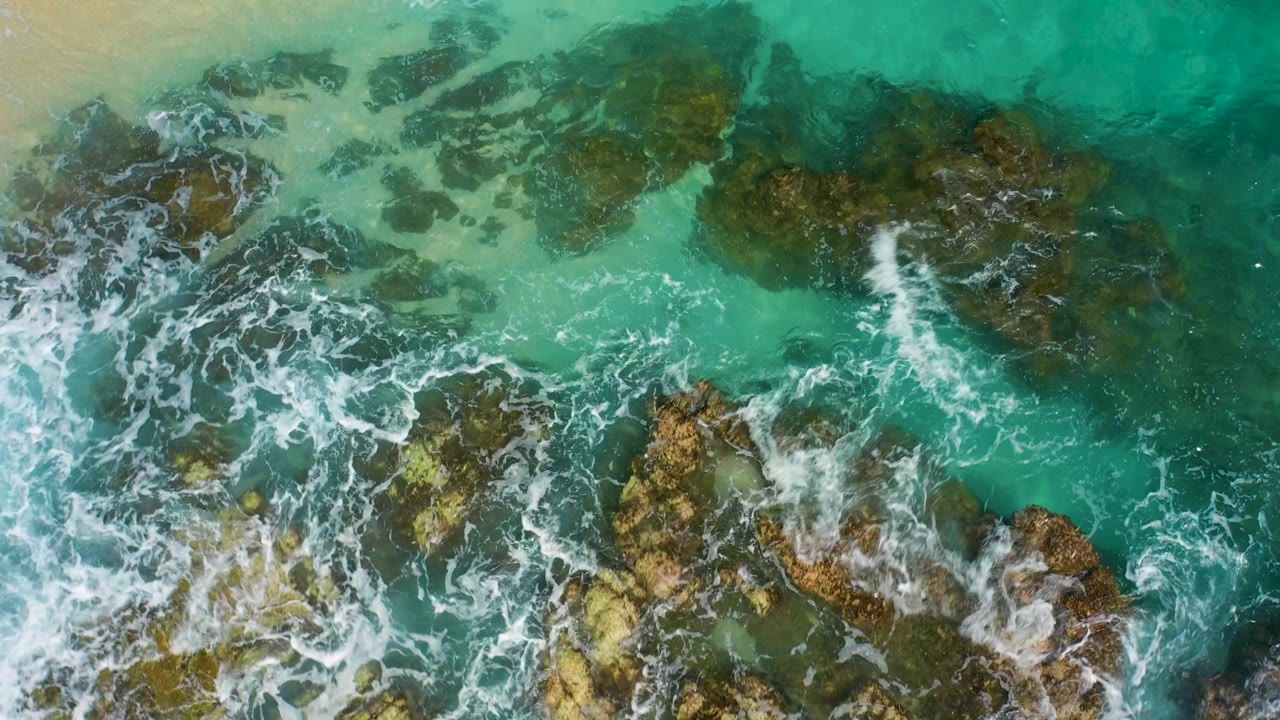 Top-down frame from Desert Park Natural Reserve, Baja California Sur, where foam pushes in from the left across jagged brown rocks and clear green-blue water, with no visible people.