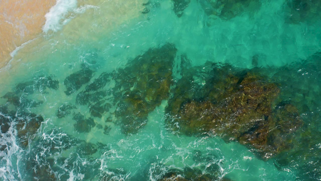 Top-down aerial view in Desert Park Natural Reserve, Baja California Sur, showing turquoise water, white foam, dark submerged rocks, and a strip of sand with no visible people.