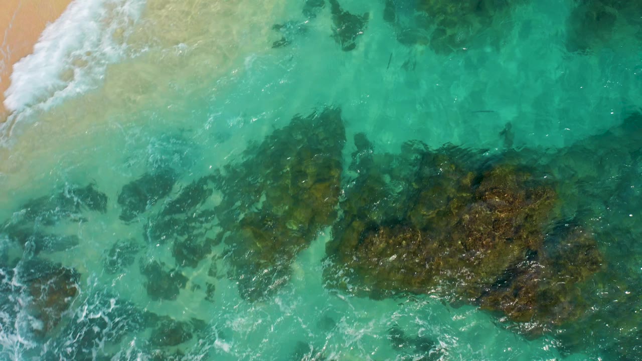 Top-down coastal frame in Baja California Sur showing white surf foam at the sandy edge and a broad dark rock patch beneath transparent turquoise water, with no visible people.