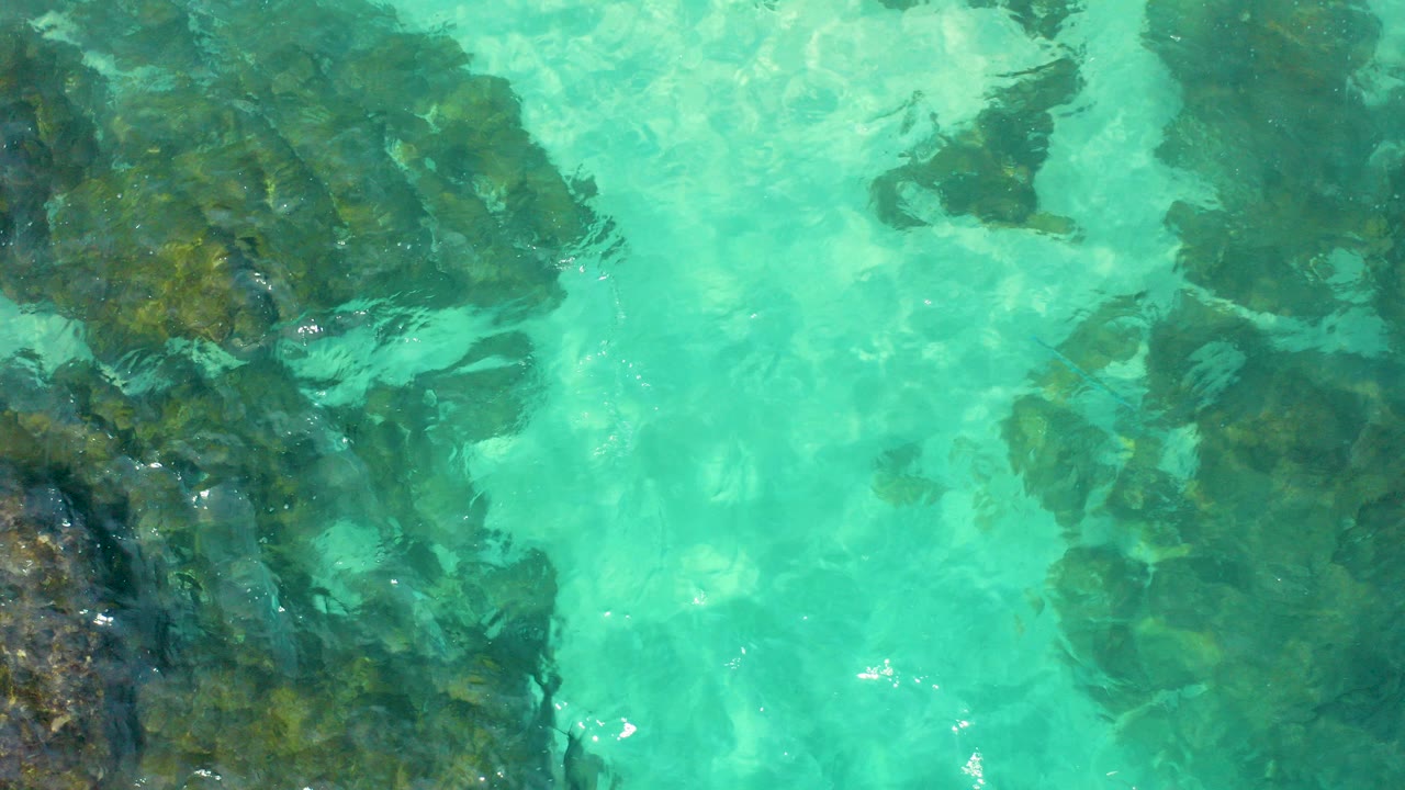 Top-down aerial frame in Baja California Sur showing turquoise water over dark submerged rocks, with small surface ripples and no visible people.
