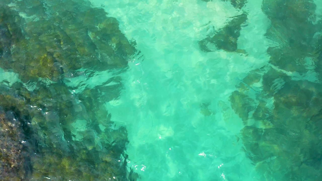 Aerial view in Baja California Sur with clear aquamarine water crossing over dark green-brown rock patches and no visible people.