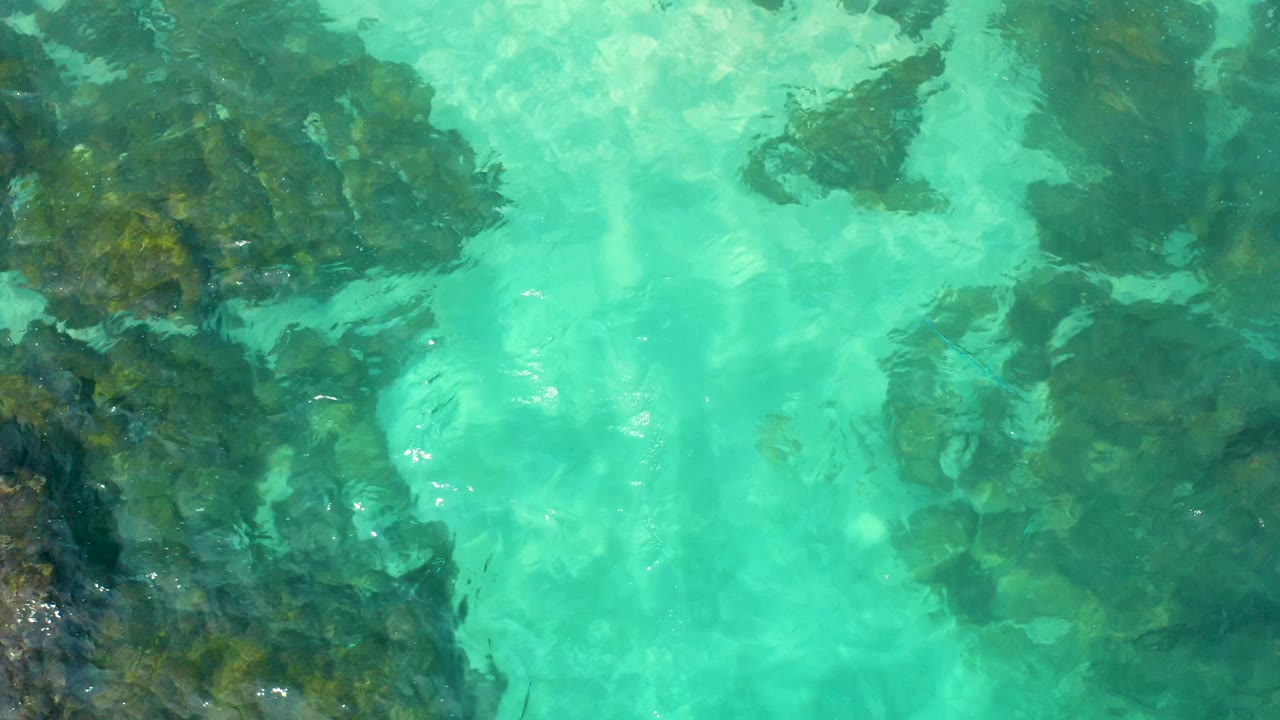 Overhead frame in Baja California Sur showing clustered underwater rocks under turquoise water, with shifting highlights and no visible people.