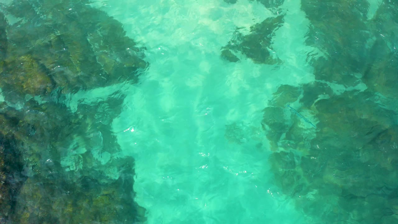 Top-down Baja California Sur water view with dark submerged rocks along the left edge, pale turquoise open water, and no visible people.