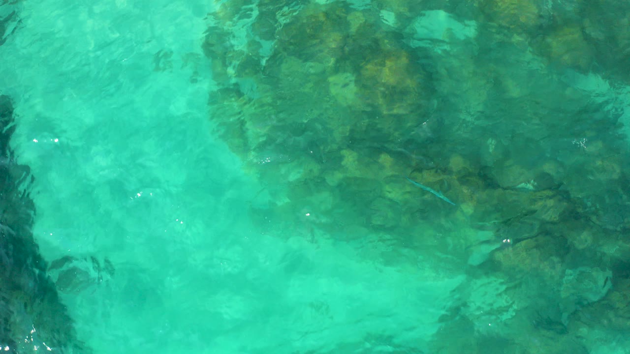 Overhead view of turquoise water in Baja California Sur with dark submerged rocks below soft surface ripples and no visible people.