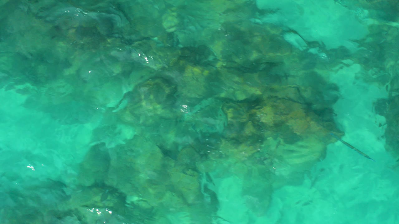 Top-down aerial frame from Desert Park Natural Reserve in Baja California Sur showing turquoise water over dark submerged rocks, with no visible people.