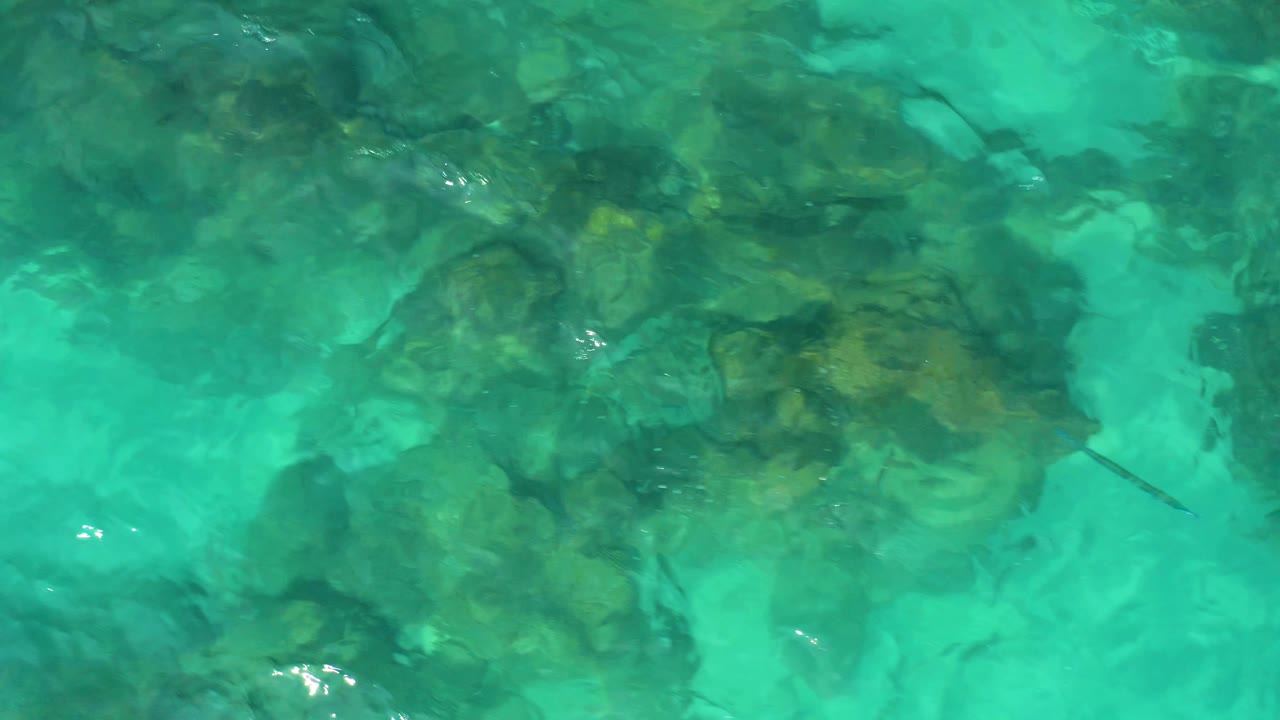 Aerial overhead view in Baja California Sur with clear turquoise water, clustered rocks beneath the surface, and no visible people.