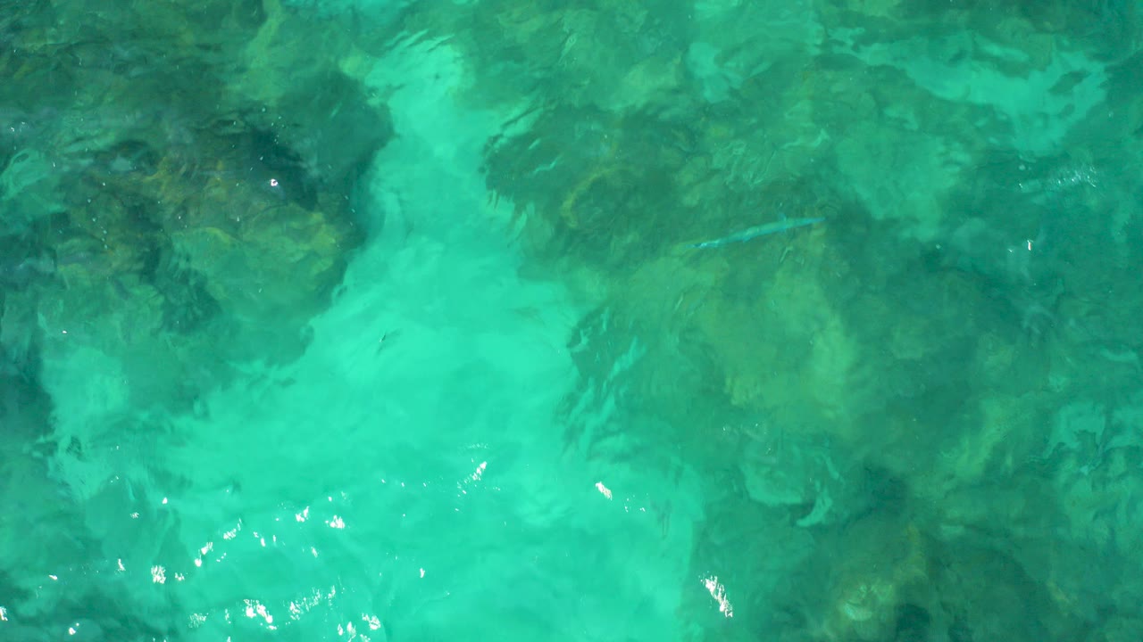 Top-down aerial view of turquoise shallow water in Baja California Sur, Mexico, with dark submerged rocks under rippled surface water and no visible people.