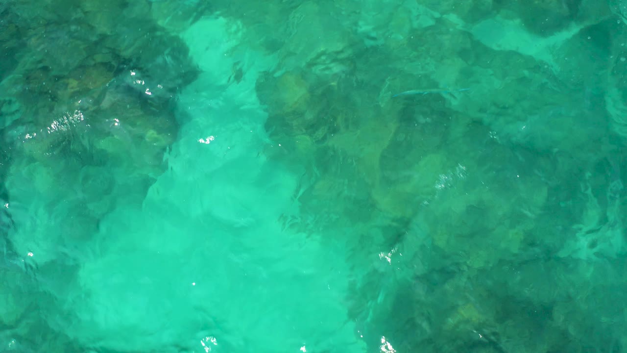 Aerial top-down view in Baja California Sur, Mexico, with turquoise ripples crossing over a mottled rocky seabed and no visible people.