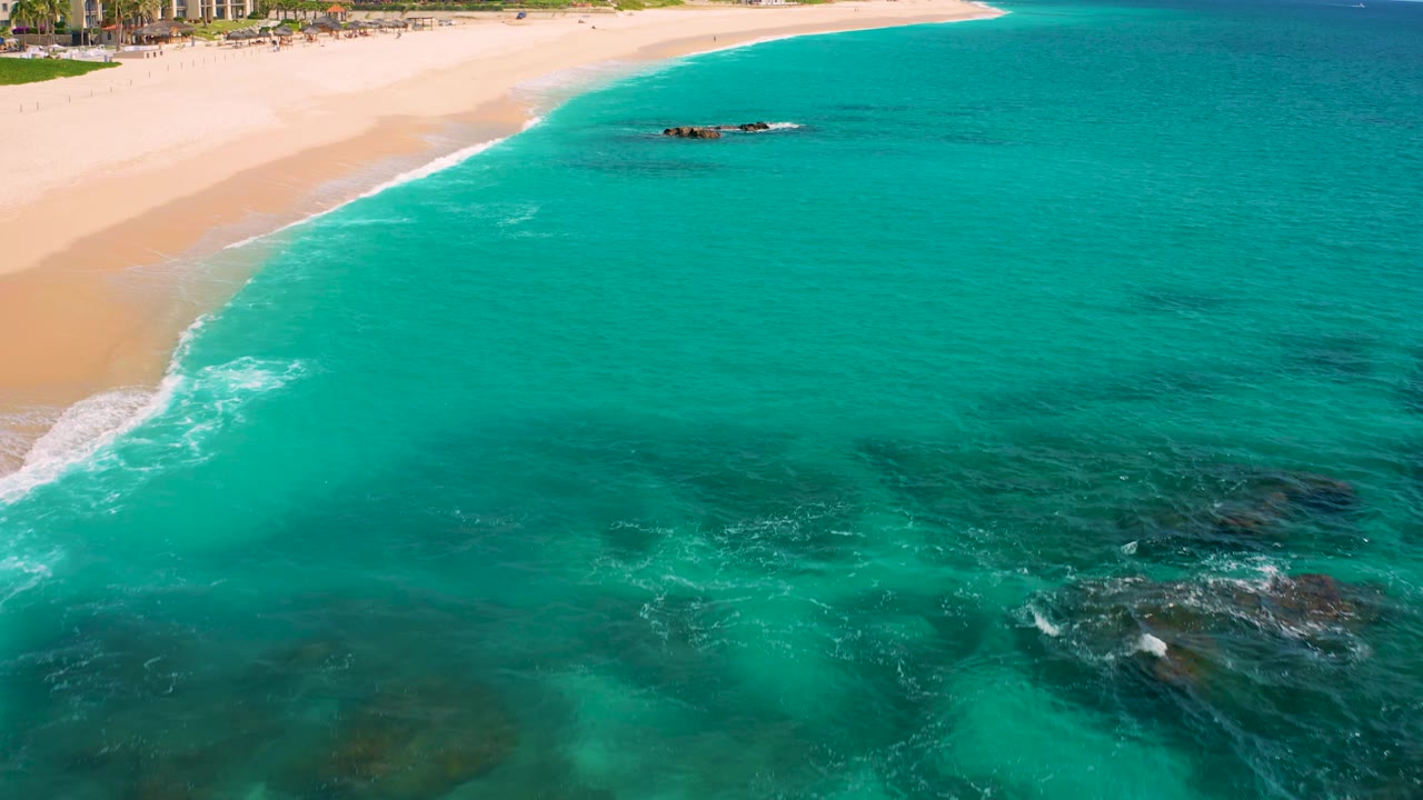 Aerial view at Desert Park Natural Reserve with broad turquoise water in the foreground, dark submerged rock patches to the right, a sandy beach at the top, and no visible people.