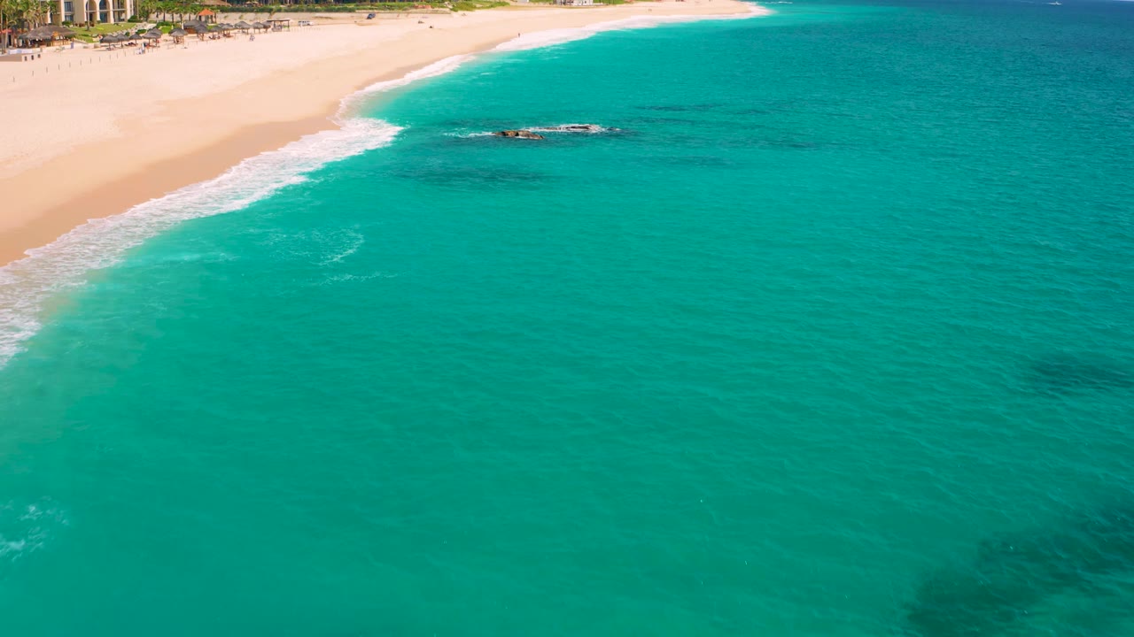 Aerial view over the Baja California Sur shoreline, with the rock outcrop slightly right of center, white foam meeting the sand, resort structures at the top edge, and no visible people.
