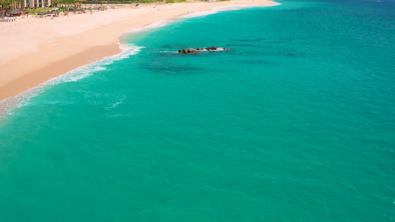 Baja California Sur coastline seen from a drone angle, with the sandy beach cutting diagonally across the upper frame, the rock cluster near the center, a small curl of white foam below, and no visible people.