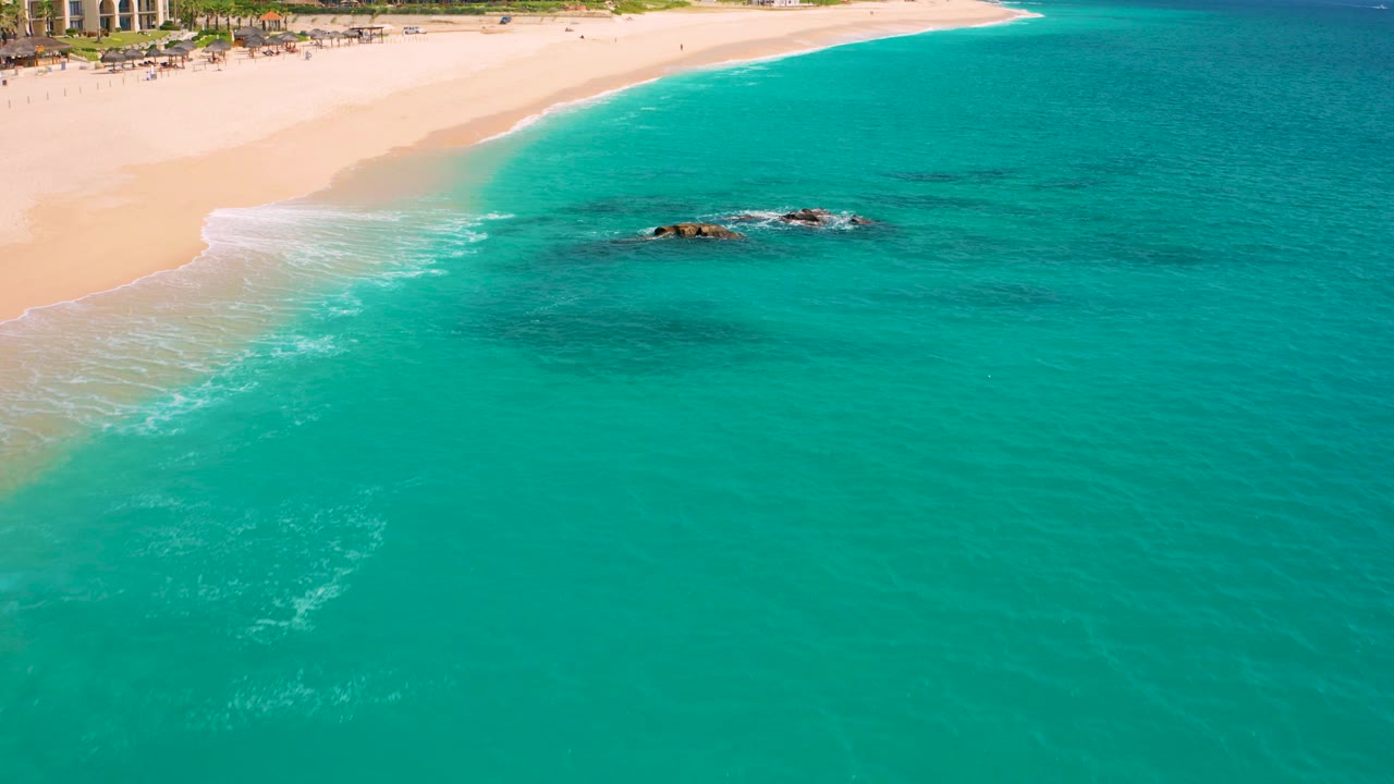 Aerial frame from Desert Park Natural Reserve in Baja California Sur showing turquoise water, pale sand, dark rocks near the surf, resort-style buildings at the beach edge, and no visible people.