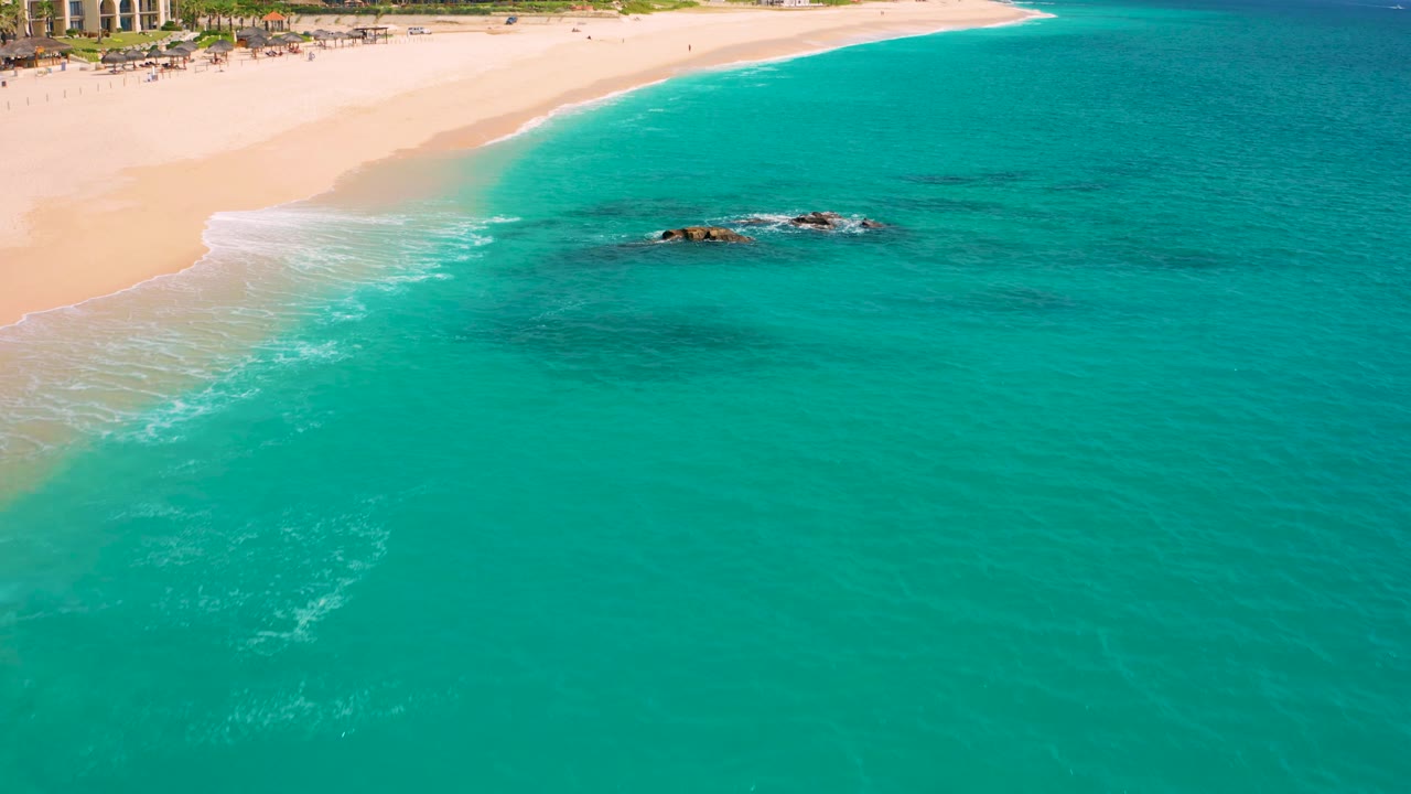 High-angle beach and turquoise water with a small rock cluster in the surf.