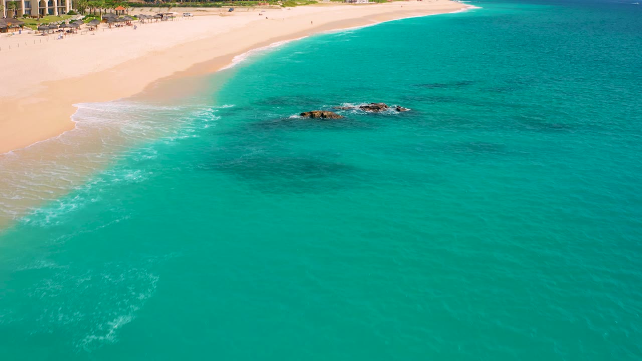 Aerial view in Baja California Sur with a curved sandy shoreline, clear turquoise water, a low group of dark rocks breaking the surface, and no visible people.