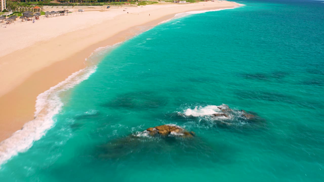 High-angle aerial frame over Desert Park Natural Reserve in Baja California Sur, Mexico, showing turquoise ocean, pale sand, dark submerged rocks, small white surf breaks, and no visible people.