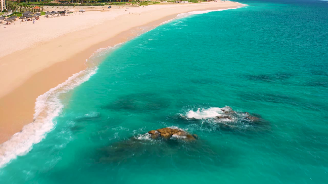 Aerial view at Desert Park Natural Reserve in Baja California Sur, Mexico, with the surf line curving along pale sand and dark submerged rocks sitting below the turquoise surface, no visible people.