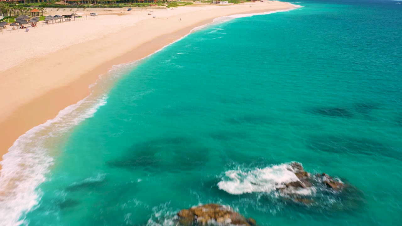 Drone frame over Desert Park Natural Reserve in Baja California Sur, Mexico, showing more open turquoise water, a diagonal beach line, foamy surf over rocks near the lower right, and no visible people.