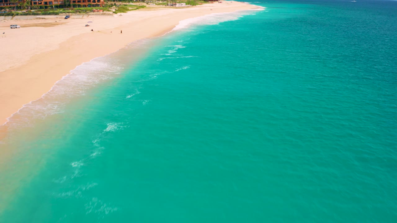 Aerial view in Baja California Sur with turquoise water dominating the frame, a pale beach along the upper left, low surf at the shoreline, resort-style buildings in the background, and no clearly visible people.