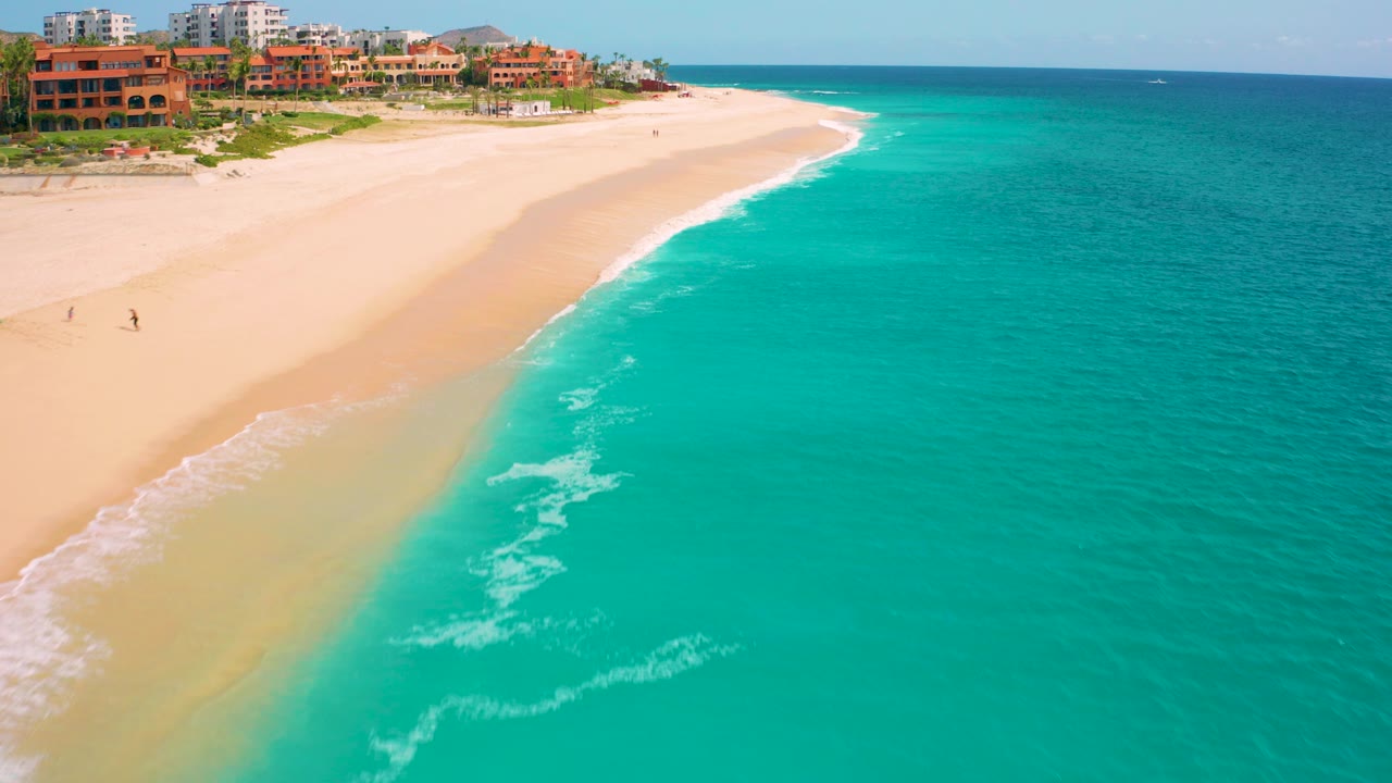 Aerial view of turquoise water, sandy beach, surf, and resort buildings.