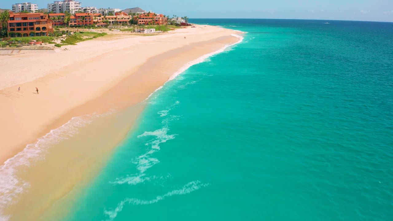 Aerial view at Desert Park Natural Reserve in Baja California Sur, Mexico, where a diagonal strip of surf separates bright turquoise water from a wide sandy beach, with resort buildings along the upper shore and no clearly visible people.