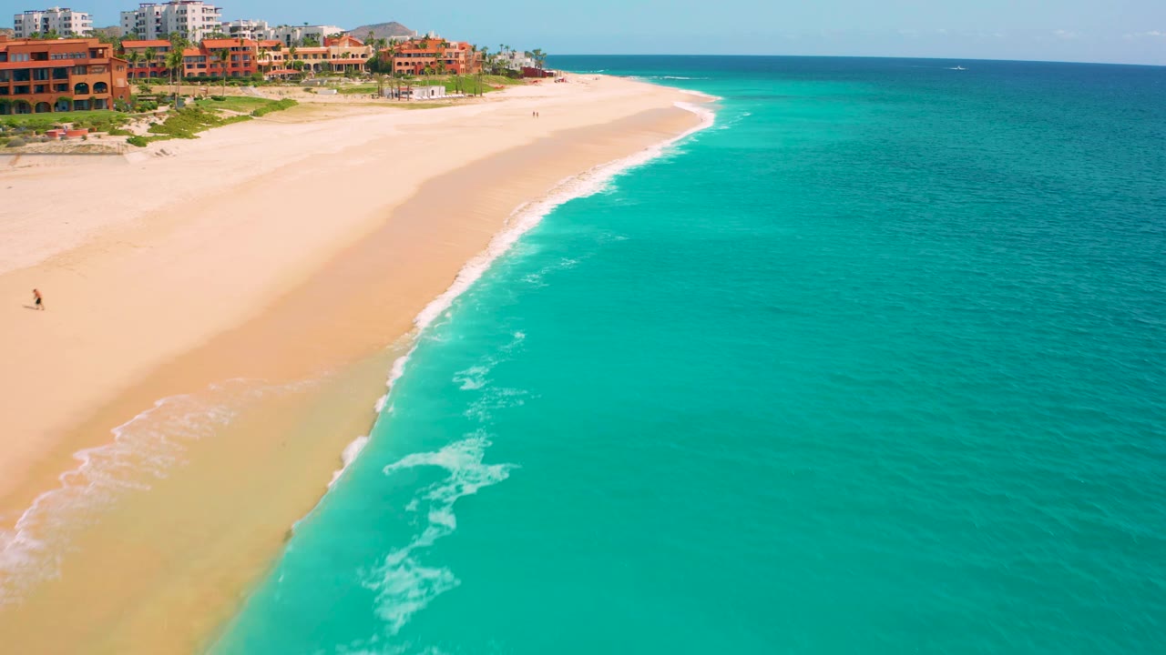 Drone-height frame over the Baja California Sur coast showing more open turquoise water to the right, a curving sandy beach to the left, surf foam near the center, resort buildings at the top edge, and no clearly visible people.