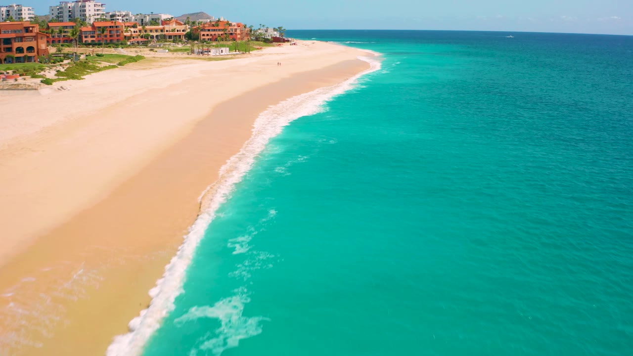 High-angle frame from Desert Park Natural Reserve, Baja California Sur, Mexico, with a white wave line running along the beach, turquoise water across the right half, terracotta buildings near the upper left, and no clearly visible people.