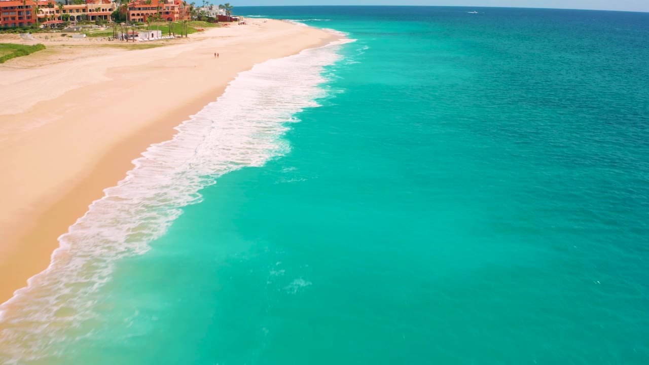 Aerial view of Desert Park Natural Reserve in Baja California Sur, Mexico, where the surf line sits higher in frame and clear turquoise water spreads across the right side with tiny people visible on the sand.