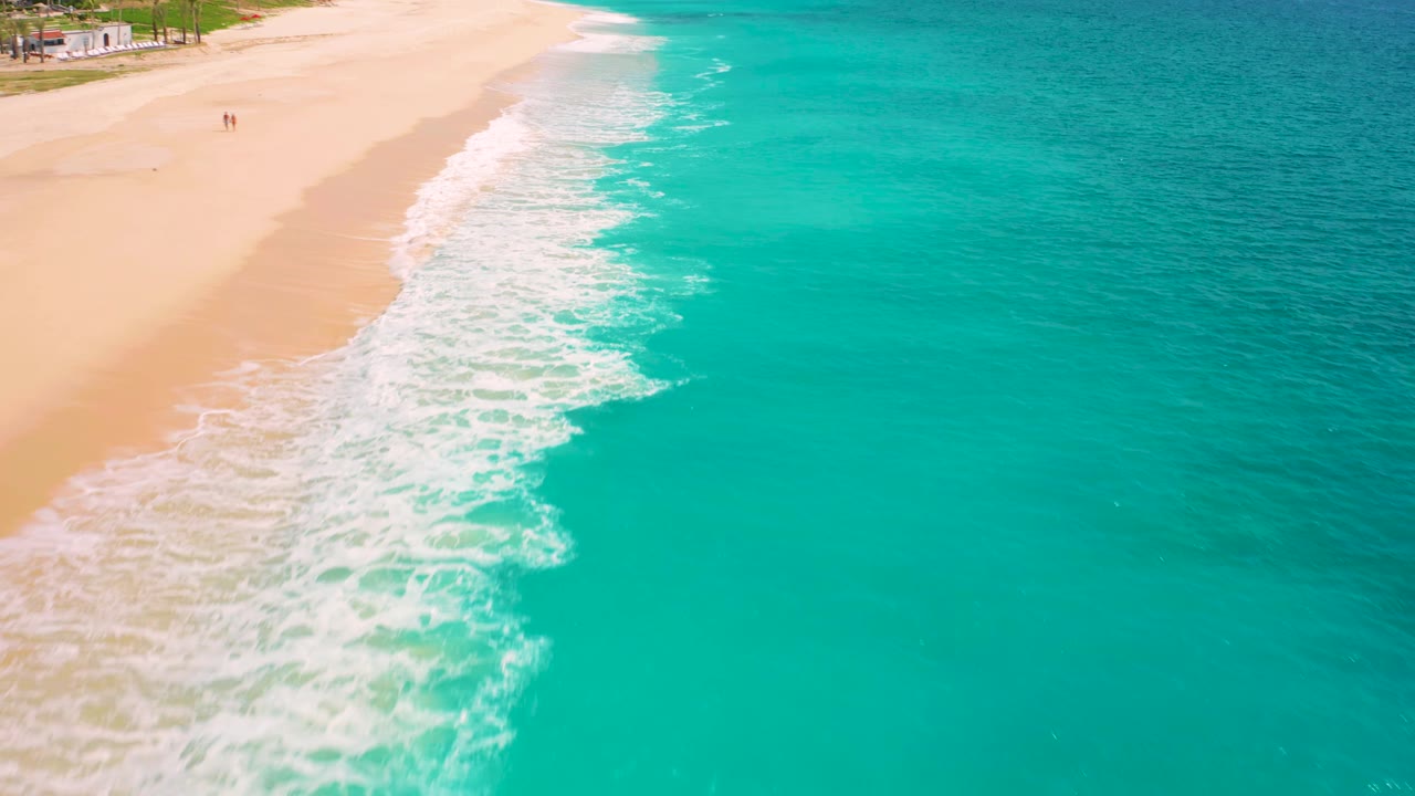 High aerial frame in Baja California Sur, Mexico, with a diagonal band of white foam between tan beach and turquoise water, plus tiny distant people near the sand.
