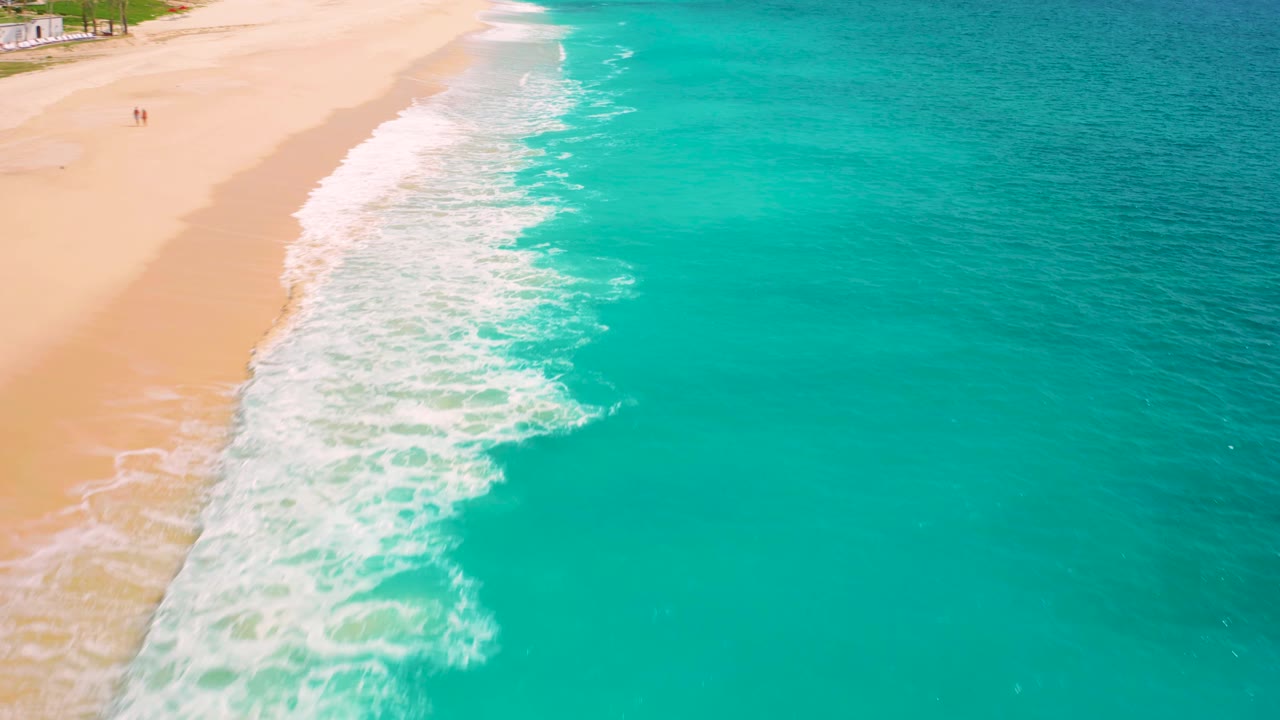 Drone-style overhead frame in Baja California Sur, Mexico, showing open teal water on the right and a narrow sandy beach edge with white surf on the left.