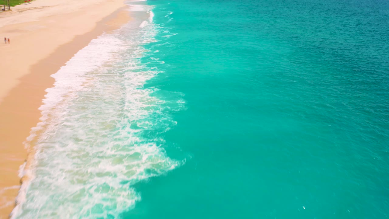 High-angle view in Baja California Sur, Mexico, with bright surf spread across wet sand, turquoise sea to the right, and tiny distant figures at the far left edge.