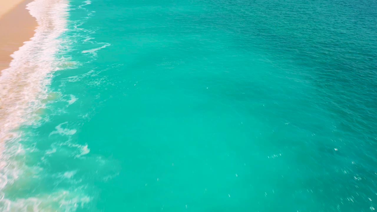 High-angle ocean frame from Baja California Sur with foamy surf brushing the left shoreline and deeper teal water spreading to the right, with no visible people.