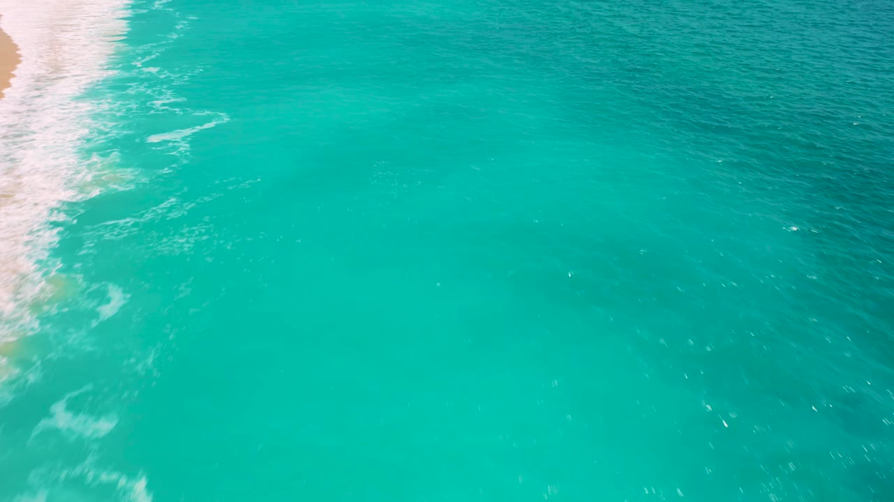 Aerial frame over Desert Park Natural Reserve water, with pale sand and white wave wash cropped at the upper left and broad turquoise sea filling the frame, with no visible people.