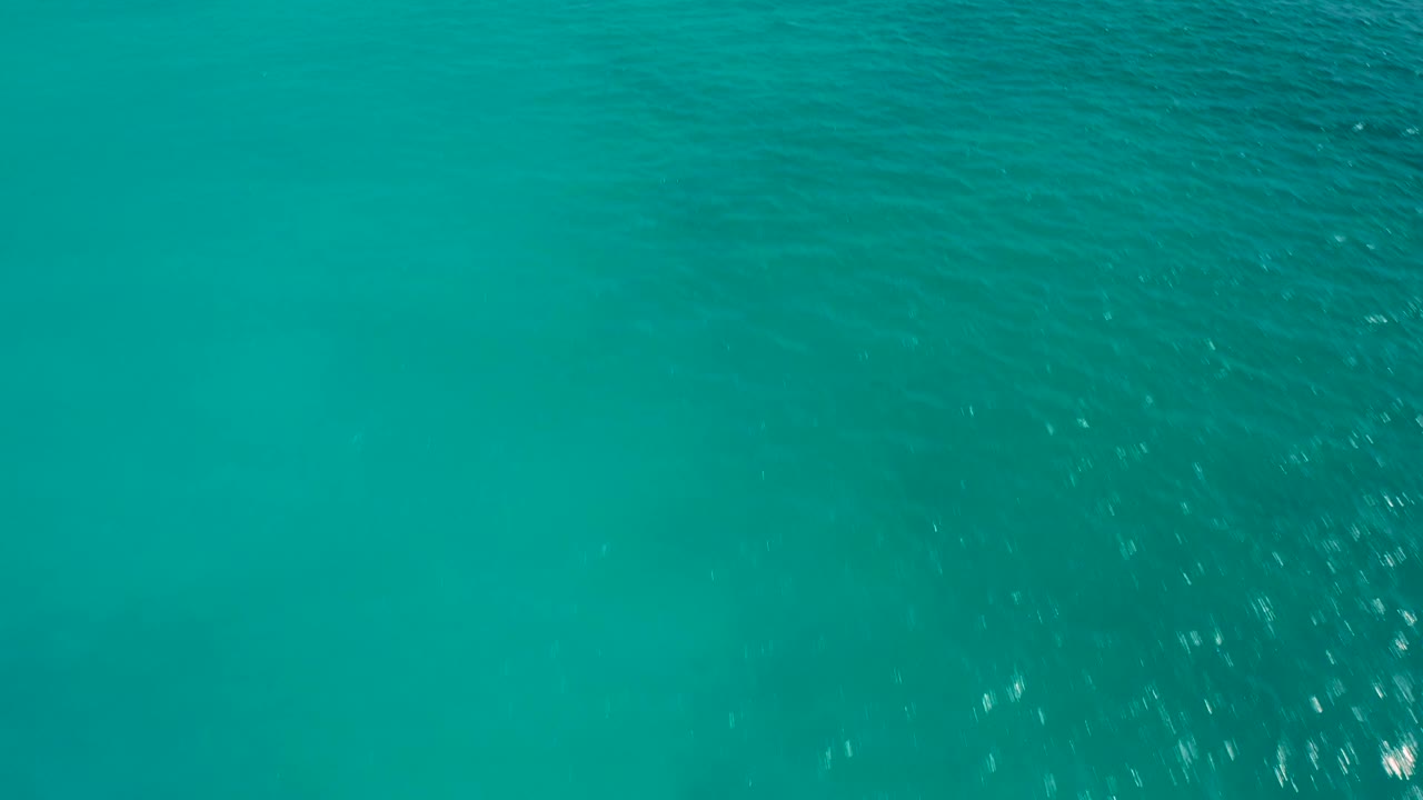 Aerial view of teal and aquamarine ocean surface with small bright reflections across the upper water texture and no visible people.