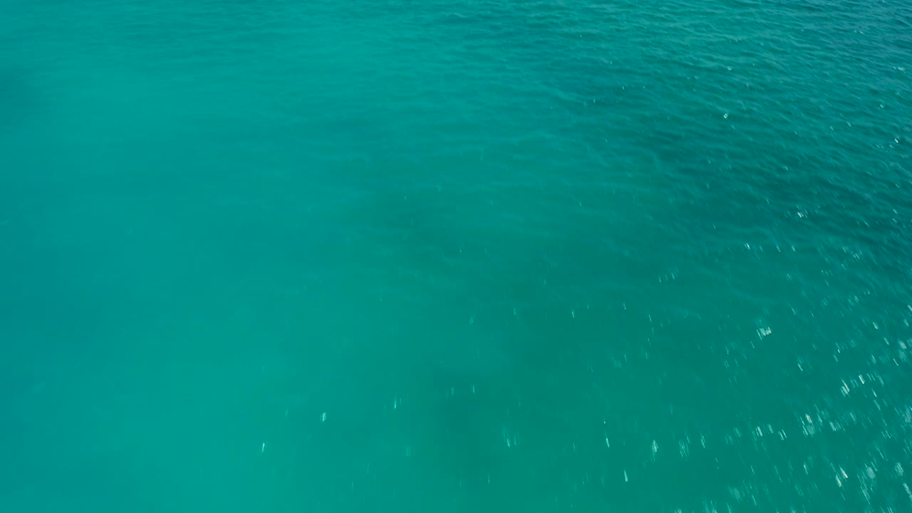 Aerial ocean frame filled by aquamarine water, low wave lines, and scattered white sparkle, with no visible people or objects.