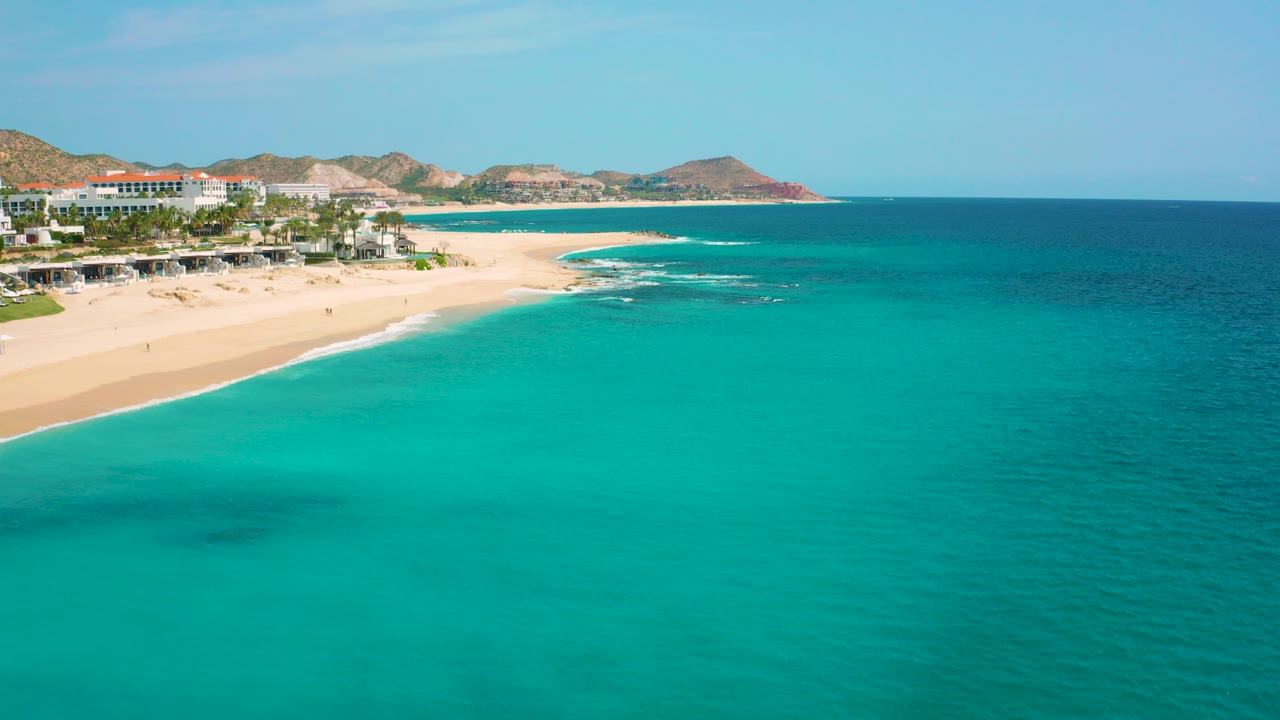 High-angle aerial frame over Desert Park Natural Reserve in Baja California Sur, showing turquoise water, pale sand, resort buildings, palms, arid hills, and no visible people.