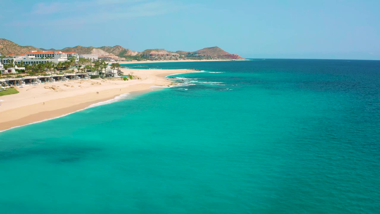 Aerial frame from Desert Park Natural Reserve with the beach curving around turquoise shallows, resort buildings on the left, hills behind, and no visible people.