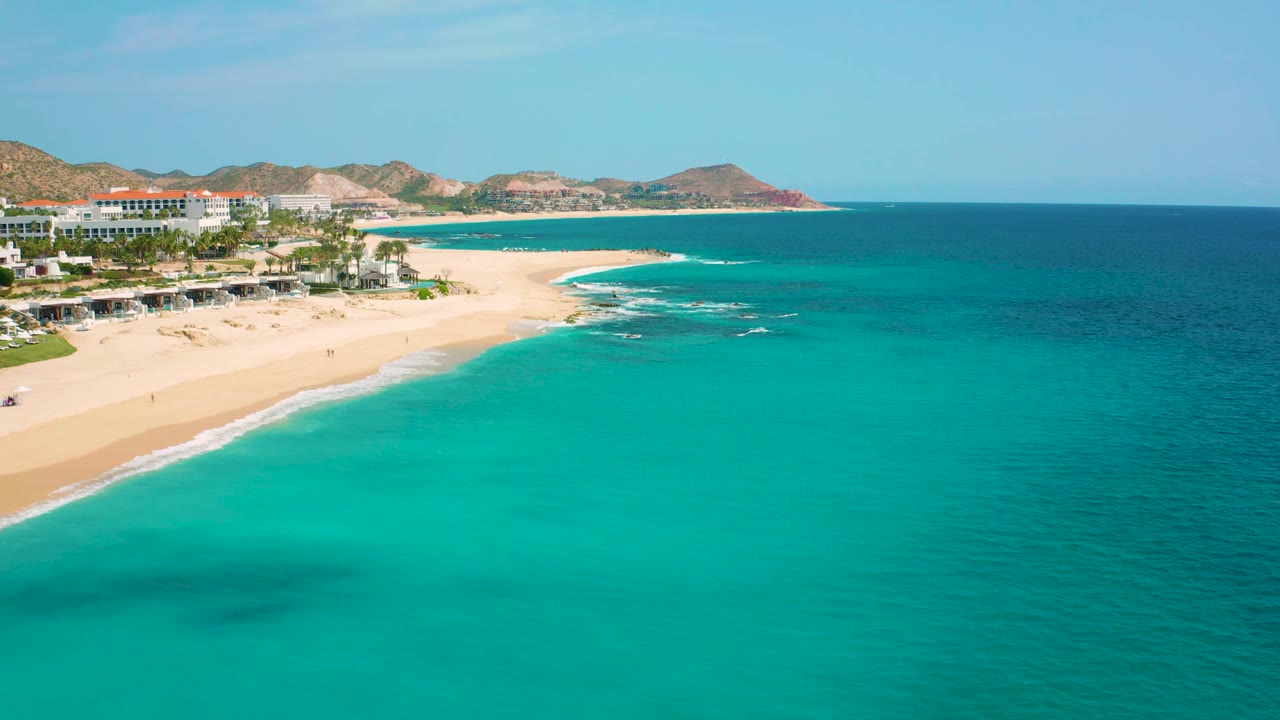 High-angle coastal frame from Desert Park Natural Reserve showing the resort area along the beach, turquoise surf water in front, arid hills across the horizon, and no visible people.