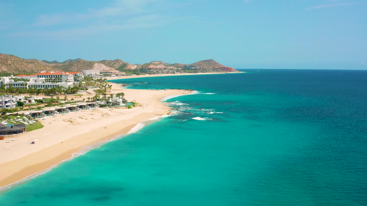 High aerial frame of Desert Park Natural Reserve in Baja California Sur, Mexico, with white resort buildings at left, pale sand below, turquoise water to the right, rocky hills in back, and no visible people.