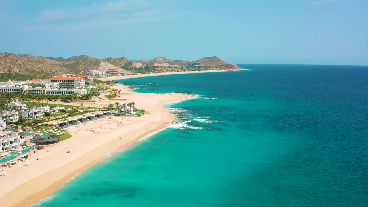 Aerial view over the Baja California Sur coastline with white buildings along the sand, clear turquoise shallows, deeper blue sea to the right, arid hills inland, and no visible people.
