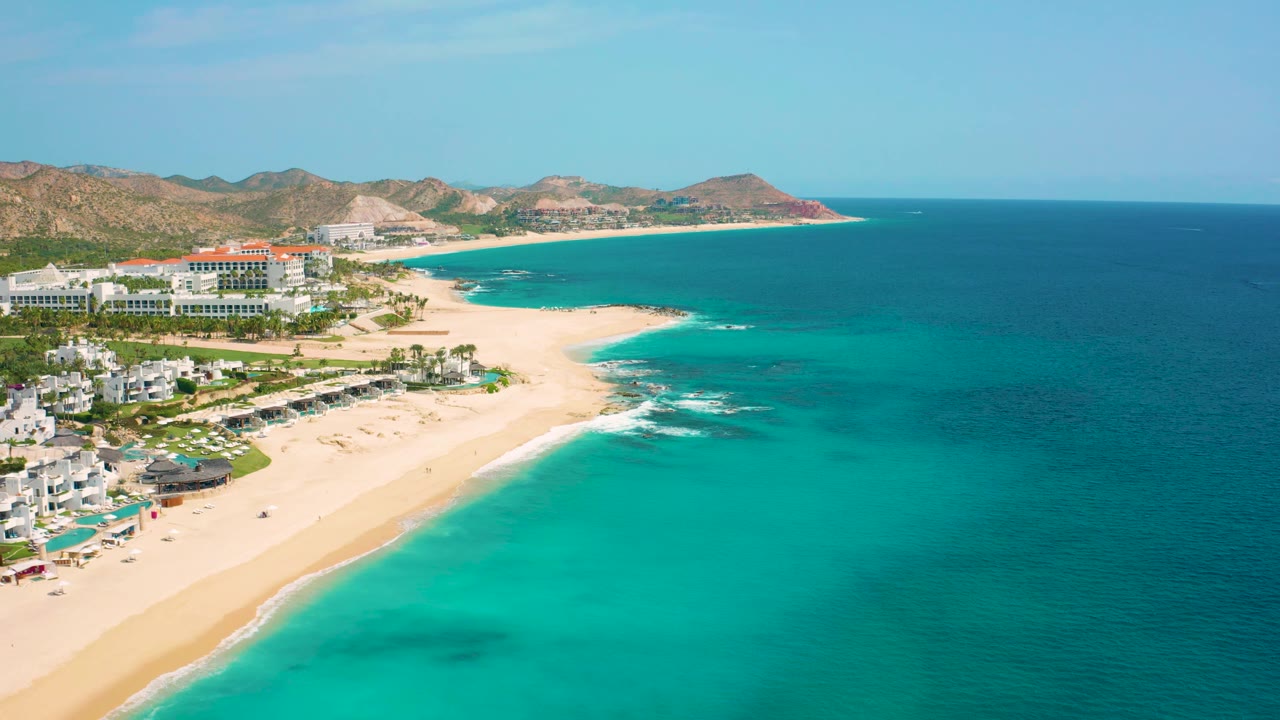 Aerial coastline with white buildings, sandy shore, turquoise sea, and distant hills.