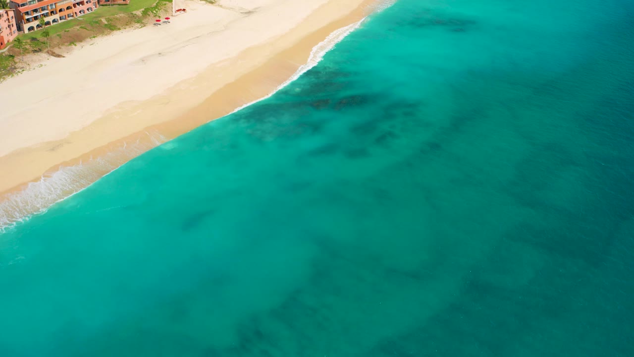 High aerial view in Desert Park Natural Reserve, Baja California Sur, with teal water filling the frame, pale beach at the top, dark underwater patches, and no visible people.