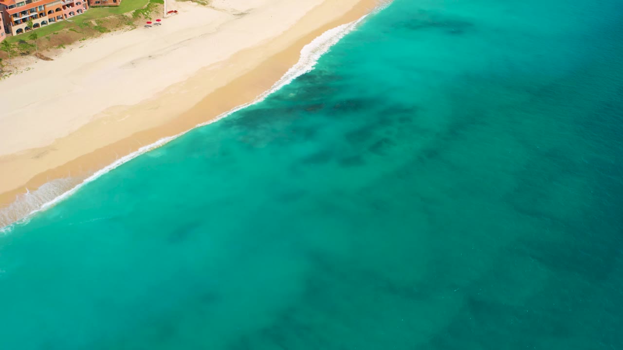High-angle frame in Desert Park Natural Reserve, Baja California Sur, with the sandy beach crossing the upper left, open teal ocean below, soft foam at shore, and no visible people.